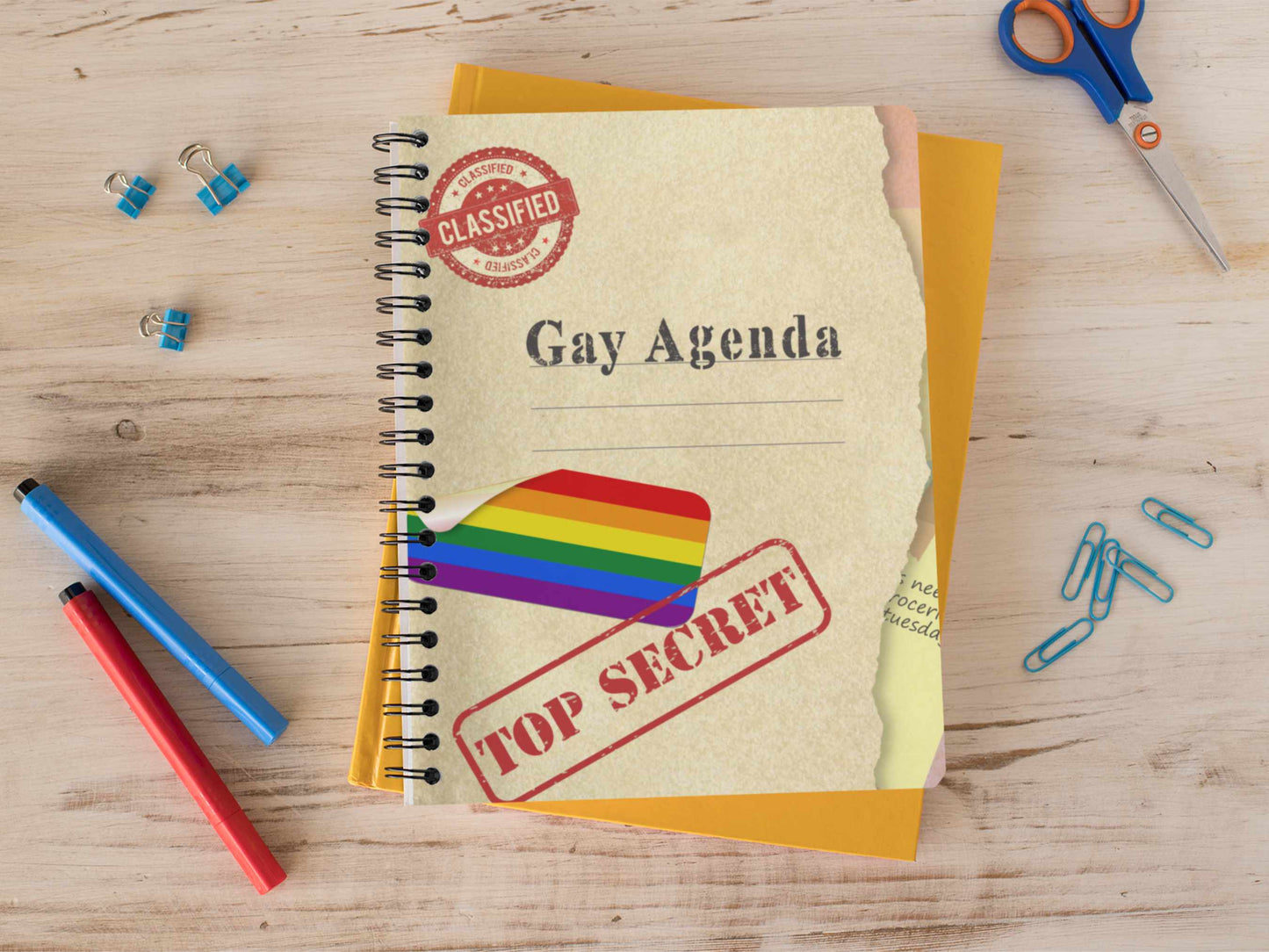 Gay Agenda spiral notebook on wooden desk surrounded by pens, scissors, and office supplies with rainbow pride cover