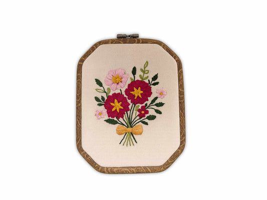 Handmade embroidery hoop art featuring pink and burgundy wildflowers with green leaves tied with golden bow on natural linen fabric in wooden hoop