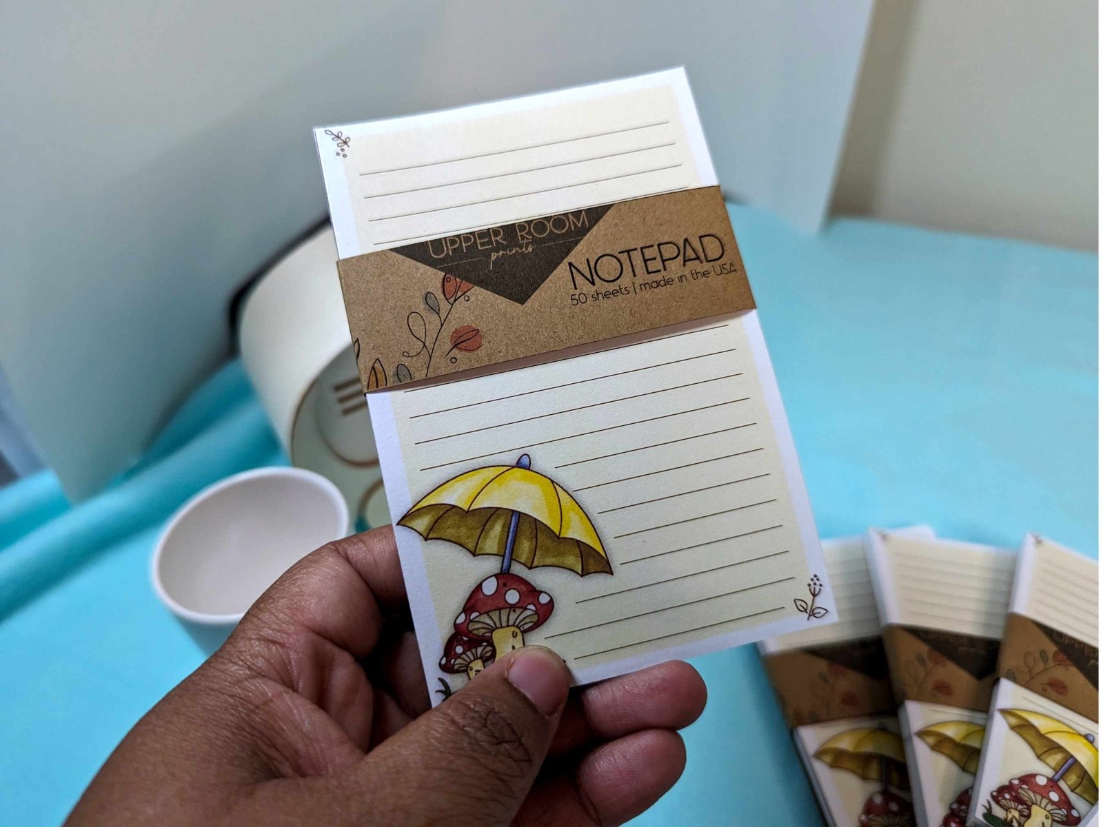 Hand holding the Mushroom Rain Notepad, showing its 4x6 size and illustrated cottagecore design.