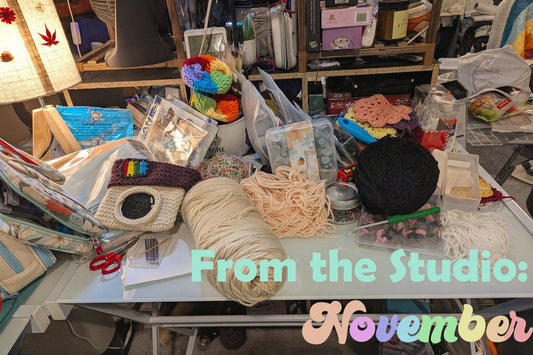 Crowded craft table in the Upper Room Prints studio covered in yarn, crochet projects, and supplies with the words “From the Studio: November” overlaid at the bottom.