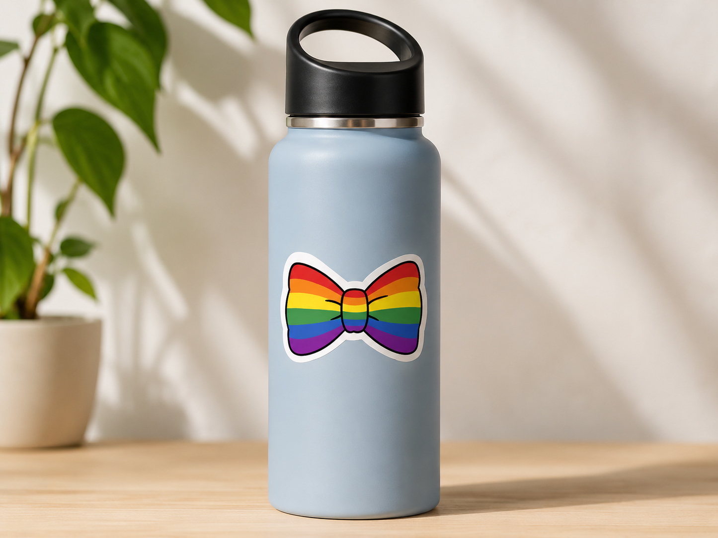 Light blue water bottle with a rainbow bow tie sticker on a wooden surface with a plant in the background.