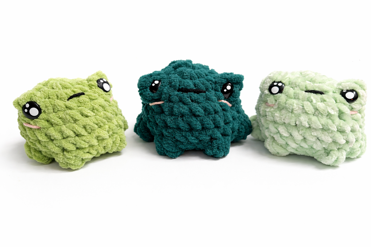 Three crochet frog plushies with cute cartoon style eyes in green shades on a white background.