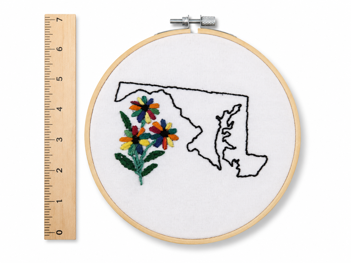 Embroidery hoop with floral design inside a map outline, next to a ruler for scale.