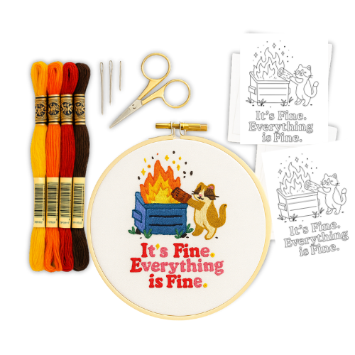 Flat lay image of the Quiet Panic embroidery kit featuring a completed “Everything is Fine” dumpster fire design in a wooden hoop, colorful thread, needles, small gold scissors, and three peel-and-stick patterns on white paper.