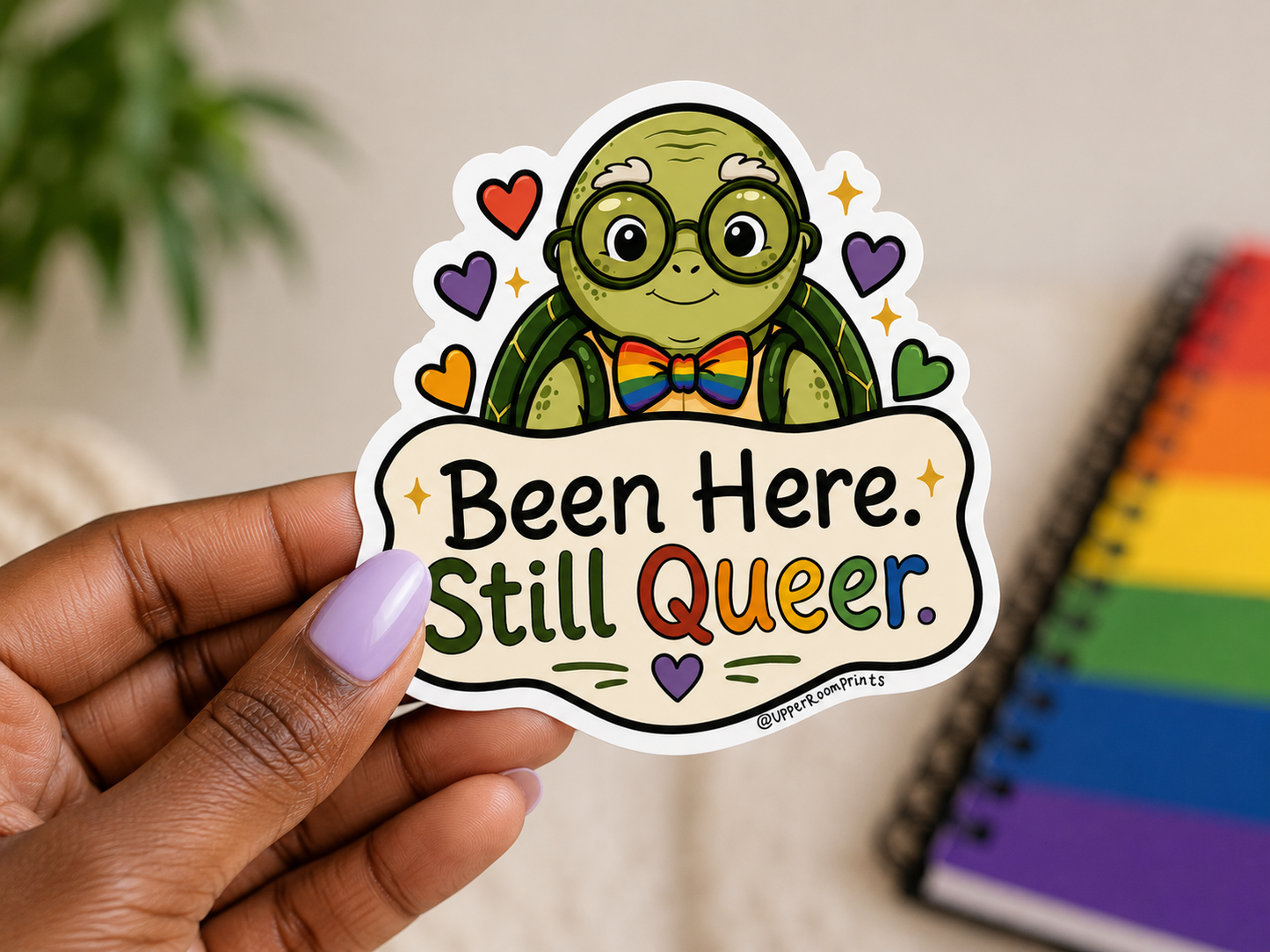 Sticker with a cartoon turtle wearing glasses and a rainbow bow tie, text 'Been Here. Still Queer.' held by a hand.