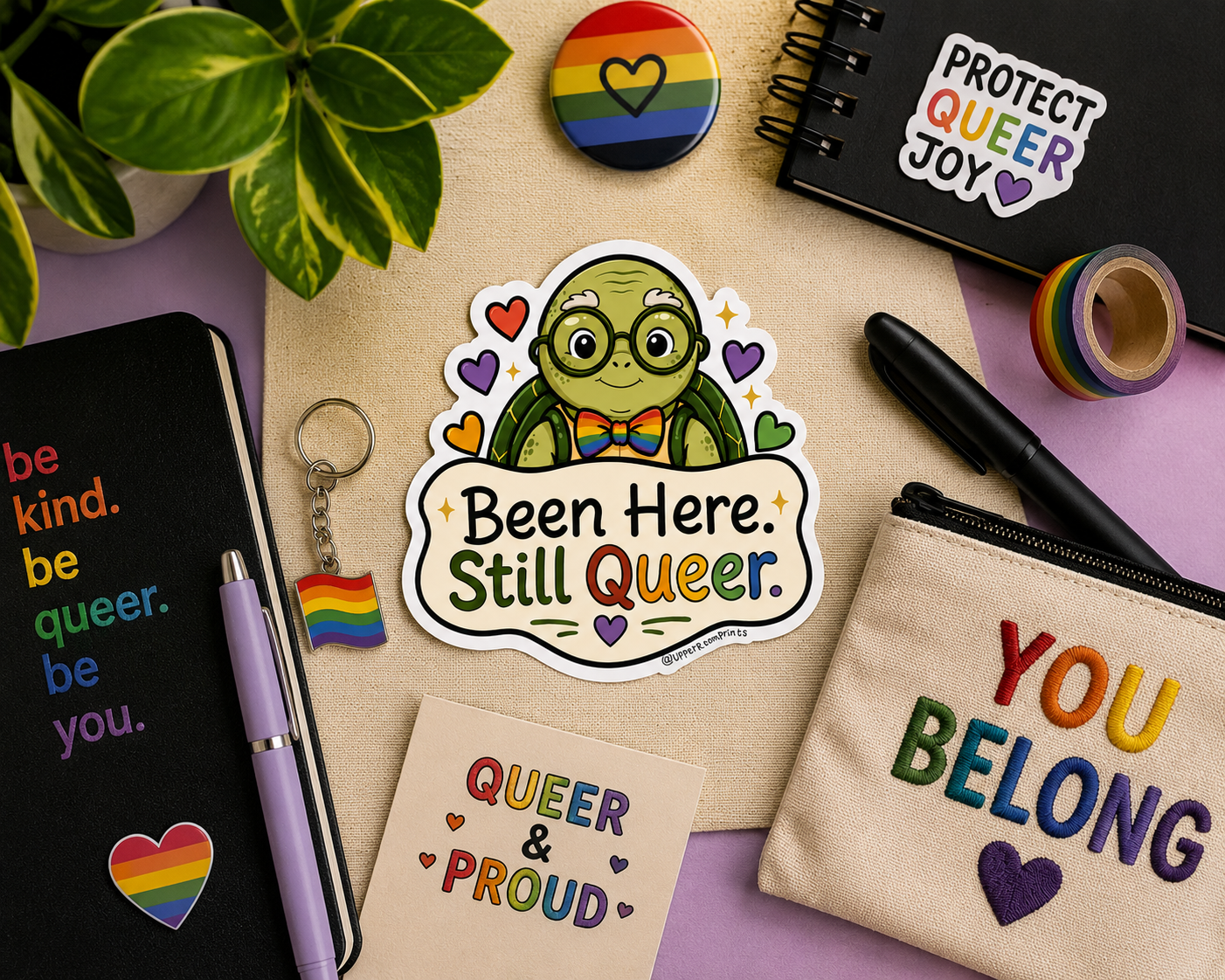 Collection of LGBTQ+ themed items including a sticker, keychain, and notebook on a wooden surface.