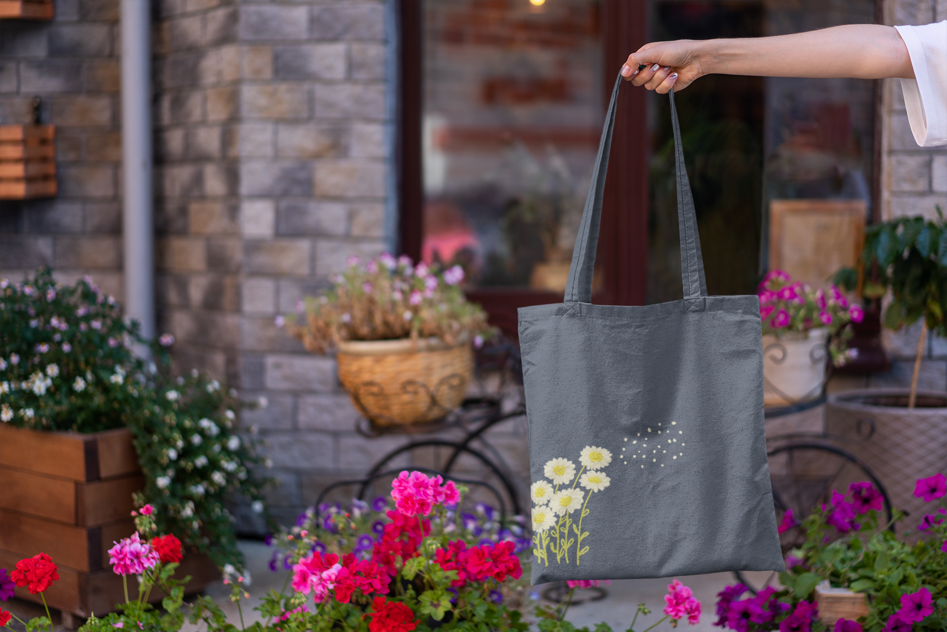 Person holding a gray tote bag with floral design in an outdoor setting with flowers and plants.