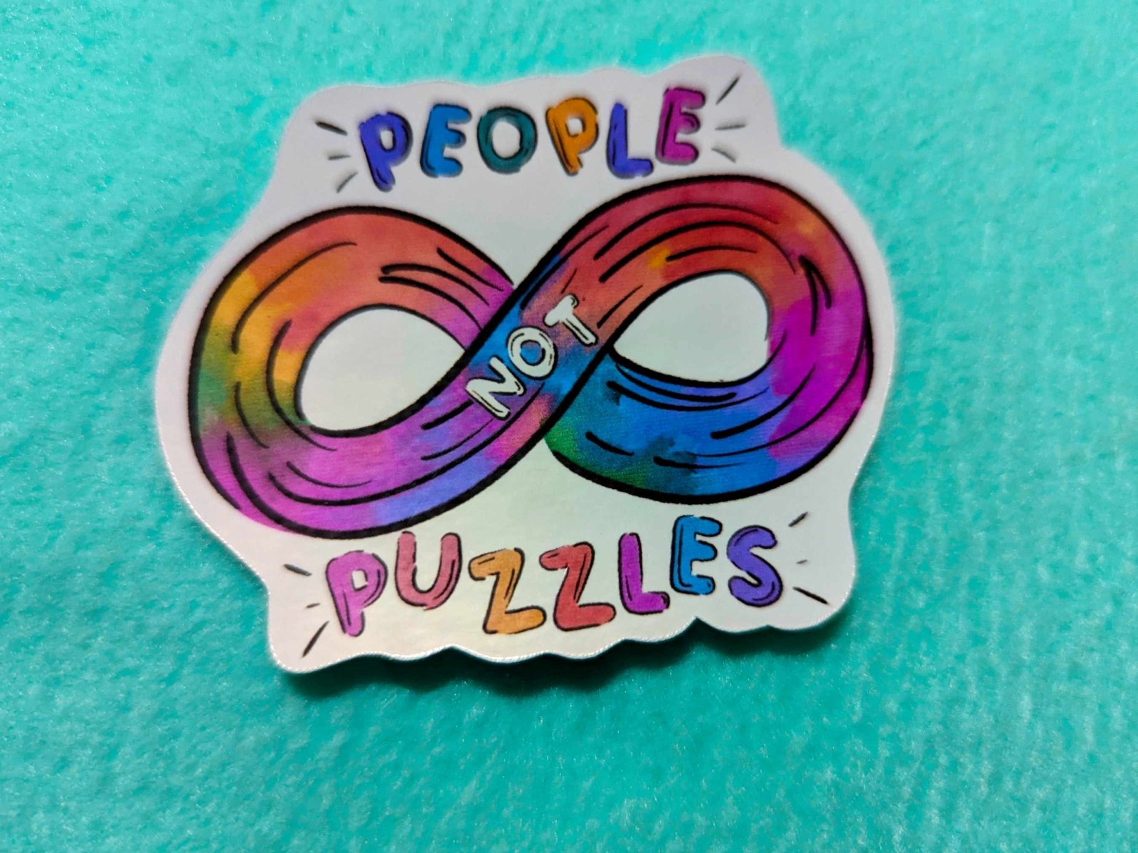 Close-up of the People Not Puzzles Sticker, showcasing its hand-drawn infinity symbol and vibrant autism pride design