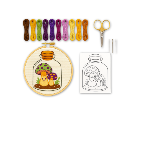 Flat lay photo of the Toadstool Terrarium embroidery kit, including a stitched mushroom design in a hoop, colorful embroidery floss, scissors, three needles, and a peel & stick pattern with a line-art version of the toadstool design.