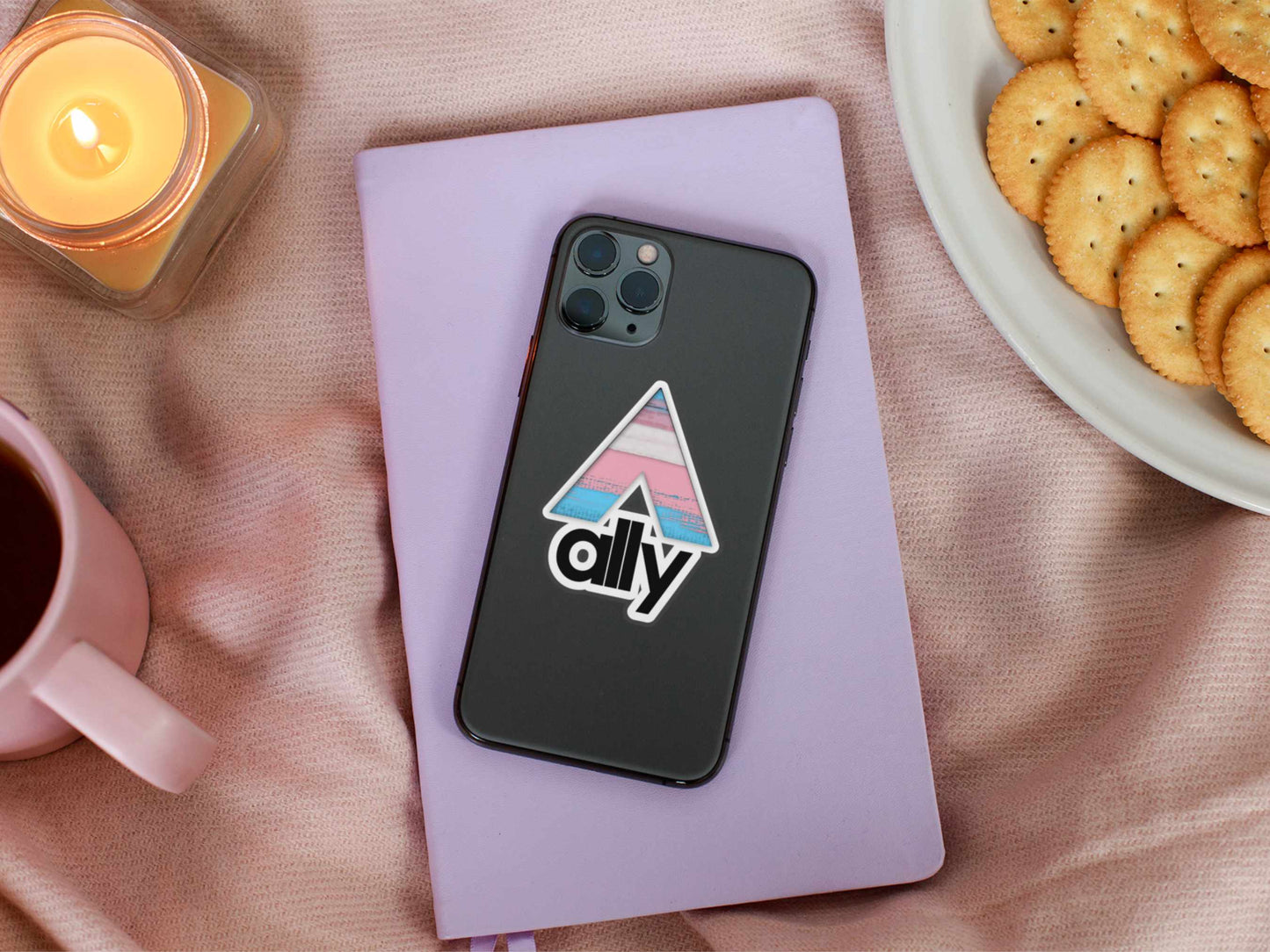 Trans flag ally sticker on phone case, resting on a pastel journal with snacks and tea nearby